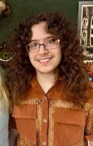 Young person with curly hair wearing glasses and a brown shirt smiling for the camera.