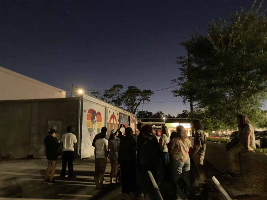 A group of people stands outside near a building at night, illuminated by artificial lighting. The building has a colorful mural on one of its walls. Trees and a fence are visible in the background.