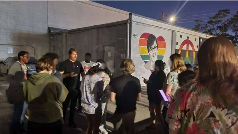 A group of people gather outdoors near a building with painted murals, including a heart with rainbow colors and a peace symbol. The setting appears to be during the evening.