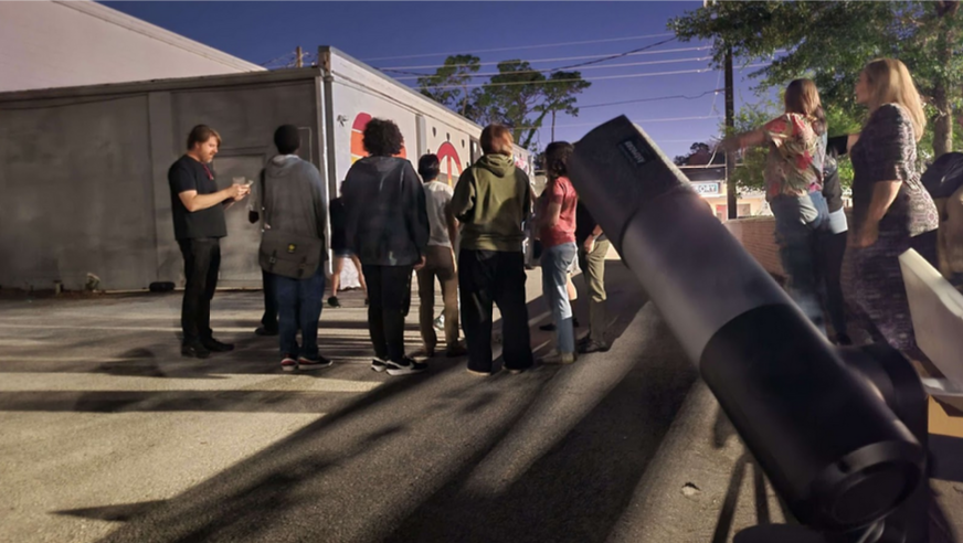 A group of people gathers near a truck and a telescope outdoors at night.