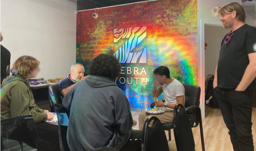 A group of people, some seated, engage in conversation in front of a colorful wall with the text "Zebra Youth." A man stands to the side with a relaxed posture.