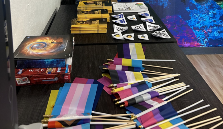 Table displaying various items including rainbow flags, brochures, booklets, and small triangular items. The flags represent different LGBTQ+ identities.