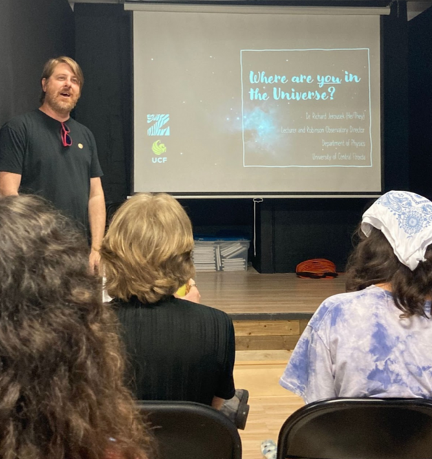 A man giving a presentation titled "Where are you in the Universe?" to an audience in a lecture room. The presentation slide shows the logo of UCF and the Department of Physics, University of Central Florida.