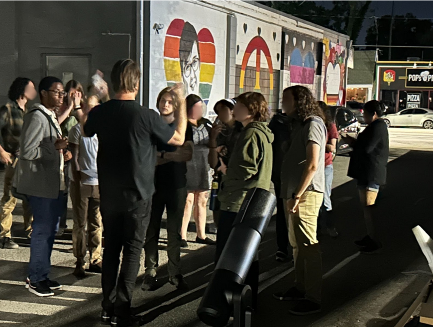 A group of people gathered on a sidewalk at night, listening to a person speaking. The background features a mural with various colorful hearts. A fast-food restaurant is visible in the distance.