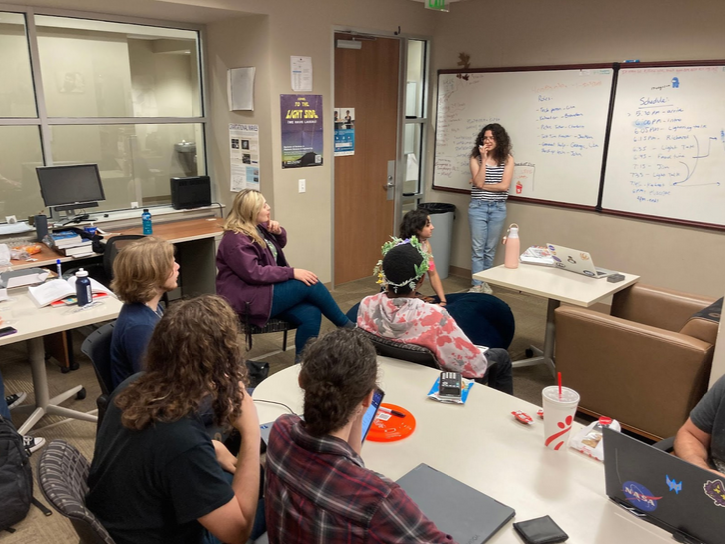 A group of people are gathered in an office, seated and listening to a person speaking in front of a whiteboard. The whiteboard contains a list of items. Various office supplies are visible.