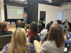 A person stands in front of a room giving a presentation titled "Big Crunch" to an audience seated and listening attentively. A projector screen displays related information and images.