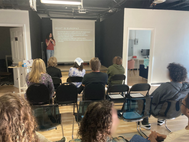 A person is giving a presentation titled "What is Europa?" to a group of seated people in a room with a projector screen. The audience is seated in rows of black chairs.