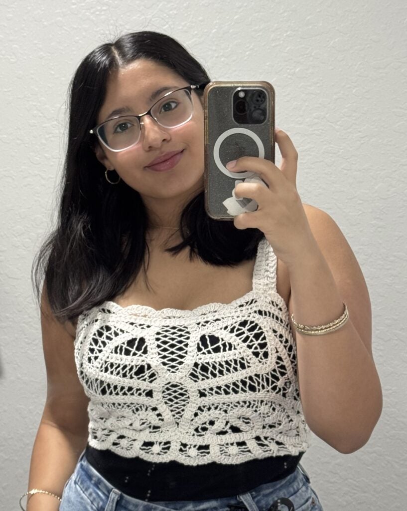 Woman with long dark hair and glasses takes a mirror selfie. She is wearing a white crochet top, blue jeans, and bracelets. The background is a plain light-colored wall.