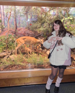 A woman stands in front of a deer and fawn museum diorama, making a heart shape with her hands and smiling at the camera.