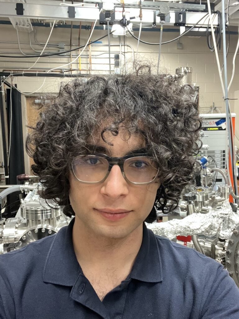 Person with curly hair and glasses taking a selfie in a laboratory setting with scientific equipment and cables in the background.