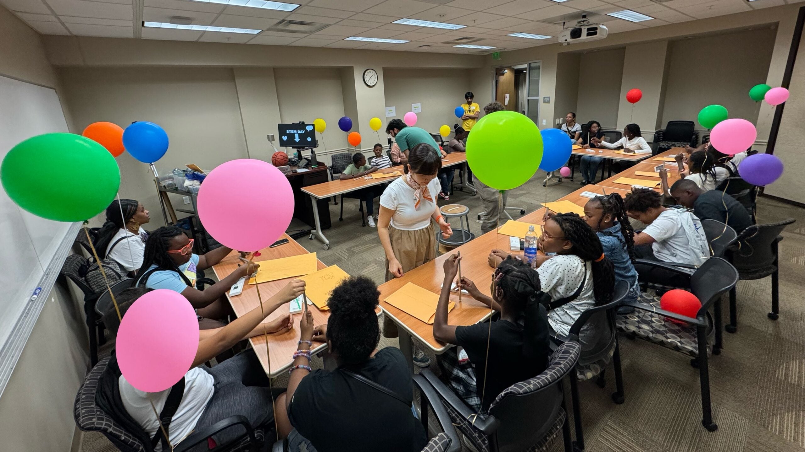 A group of people sit around tables in a conference room, working on crafts or writing, with colorful balloons attached to each chair.