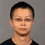 A person with short black hair, wearing glasses and a black shirt, is posed against a plain gray background.