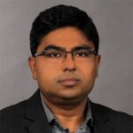 A man with short black hair, glasses, and a neutral expression, wearing a dark blazer and grey shirt, poses in front of a plain grey background.