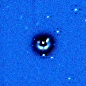 A blue-tinted astronomical image shows a central bright spot with surrounding concentric rings and scattered smaller points of light against a speckled background.