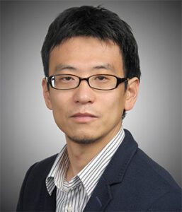 Man with short black hair and glasses wearing a dark blazer and striped shirt poses for a professional headshot against a gray background.