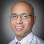 A man with glasses, a bald head, and wearing a striped shirt with a tie, smiling subtly while looking at the camera.