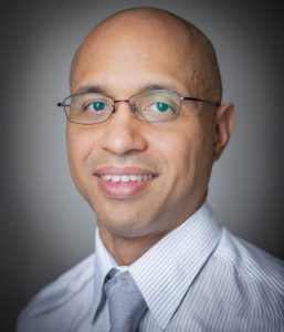 A man with glasses, a bald head, and wearing a striped shirt with a tie, smiling subtly while looking at the camera.