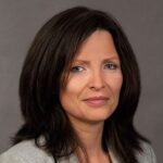 Woman with shoulder-length dark brown hair, wearing a light gray blazer and a light-colored top, posed against a plain gray background.