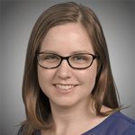 A woman with straight brown hair, wearing glasses and a blue top, smiles at the camera against a neutral background.