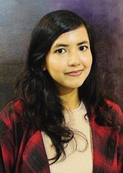 A person with long dark hair is standing in front of a textured backdrop. They are wearing a red and black plaid jacket over a light shirt.