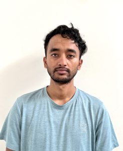 A man with short, curly hair and a beard, wearing a light blue t-shirt, stands against a plain white background, looking directly at the camera.