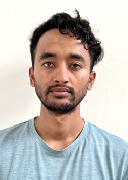 A man with short, curly hair and a beard, wearing a light blue t-shirt, stands against a plain white background, looking directly at the camera.