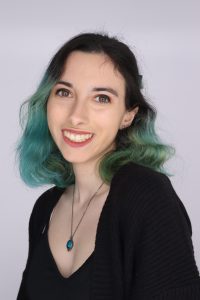 A person with shoulder-length, green-tinted hair, wearing a black top and a necklace with a teal pendant, smiles at the camera against a plain background.