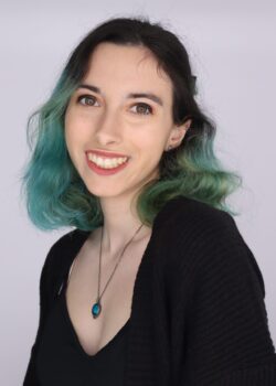 A person with shoulder-length, green-tinted hair, wearing a black top and a necklace with a teal pendant, smiles at the camera against a plain background.