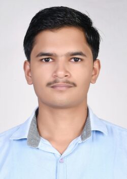 A young man with short dark hair and a mustache is wearing a light blue collared shirt and looking directly at the camera. The background is plain light gray.