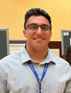 A person with short dark hair, wearing glasses and a checked shirt with a blue lanyard, stands in front of a patterned yellow wall and a bulletin board.