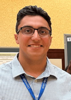 A person with short dark hair, wearing glasses and a checked shirt with a blue lanyard, stands in front of a patterned yellow wall and a bulletin board.