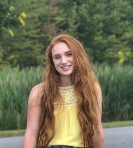 A person with long red hair, wearing a sleeveless yellow top, smiles outdoors with greenery in the background.