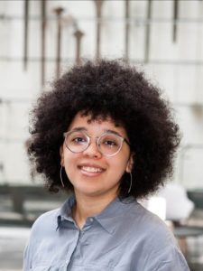 Person with curly hair, glasses, and a blue shirt smiling in an indoor setting with tools hanging in the background.