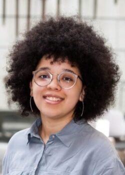 Person with curly hair, glasses, and a blue shirt smiling in an indoor setting with tools hanging in the background.