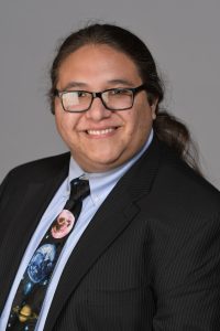 A person with glasses, long hair tied back, wearing a dark pinstripe suit, light blue shirt, and a space-themed tie, smiles at the camera against a gray backdrop.