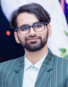 A person with glasses, a beard, and short dark hair smiles while wearing a green striped suit jacket and a light-colored shirt.