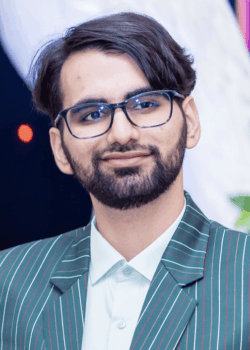 A person with glasses, a beard, and short dark hair smiles while wearing a green striped suit jacket and a light-colored shirt.