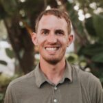 A man with short brown hair and a trimmed beard is smiling at the camera. He is wearing an olive green polo shirt and standing outdoors with greenery in the background.