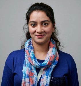 A person with a slight smile, wearing a blue top and a colorful floral scarf, in front of a plain background.