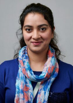 A person with a slight smile, wearing a blue top and a colorful floral scarf, in front of a plain background.