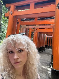 A person with curly blonde hair is taking a selfie in front of a series of bright orange Torii gates in a wooded area.