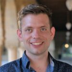 A person with short hair smiling, wearing a patterned shirt, stands outdoors with archways in the background.