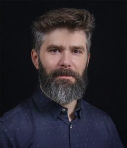 A man with short, dark hair and a full, graying beard wearing a dark blue, button-up shirt with a subtle dot pattern, against a black background.