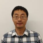 A person wearing glasses and a plaid shirt stands against a plain white background, facing the camera.