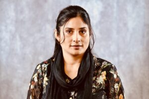 A person with long dark hair wears a floral top and black scarf, standing against a gray background.