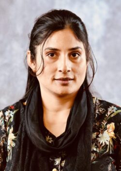 A person with long dark hair wears a floral top and black scarf, standing against a gray background.