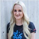 A woman with long blonde hair wearing a black "Europa Clipper" t-shirt is standing against a concrete wall, smiling at the camera.