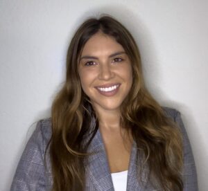 A woman with long brown hair, wearing a gray plaid blazer and a white top, stands smiling in front of a plain light-colored wall.