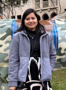 A woman stands outdoors in front of a camouflage-patterned sculpture, wearing a gray jacket and black-and-white skirt, with buildings and trees in the background.