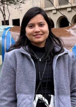 A woman stands outdoors in front of a camouflage-patterned sculpture, wearing a gray jacket and black-and-white skirt, with buildings and trees in the background.